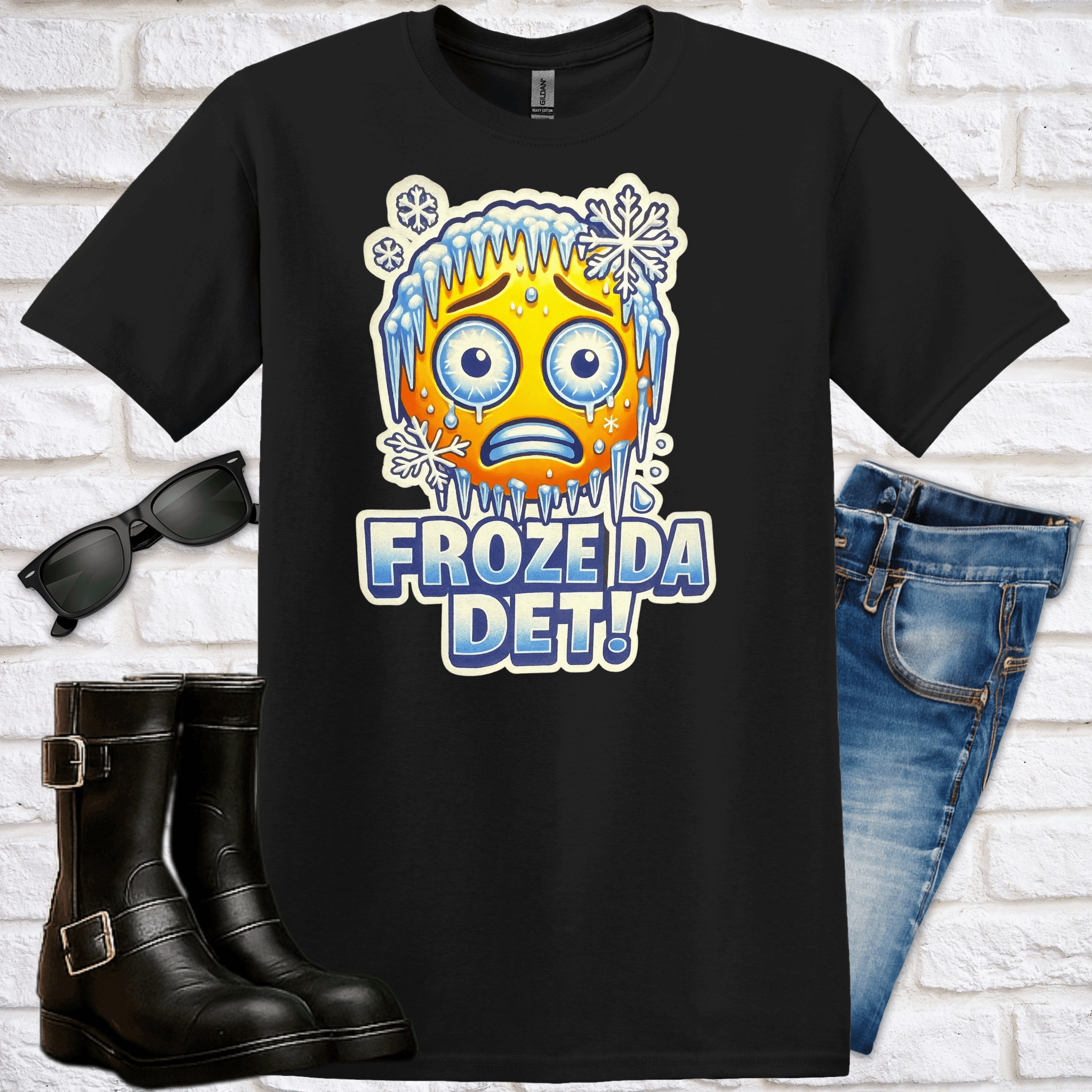Black t-shirt with a frozen emoji and text 'Froze Da Det!' on a white brick background, with sunglasses, boots, and jeans displayed around.