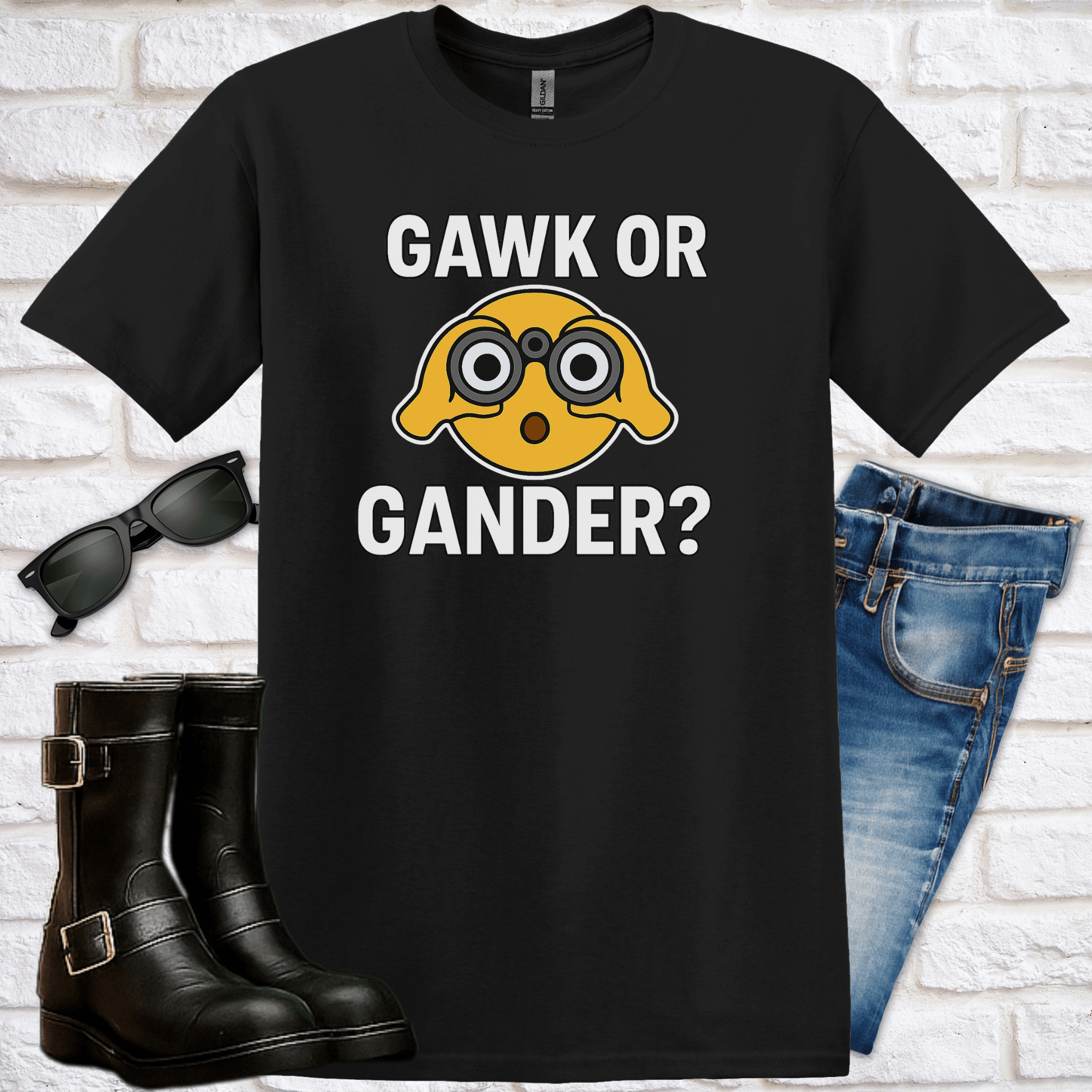 Black t-shirt with an emoji with binoculars and text saying "Gawk or Gander" with sunglasses, boots, and jeans on a white brick background