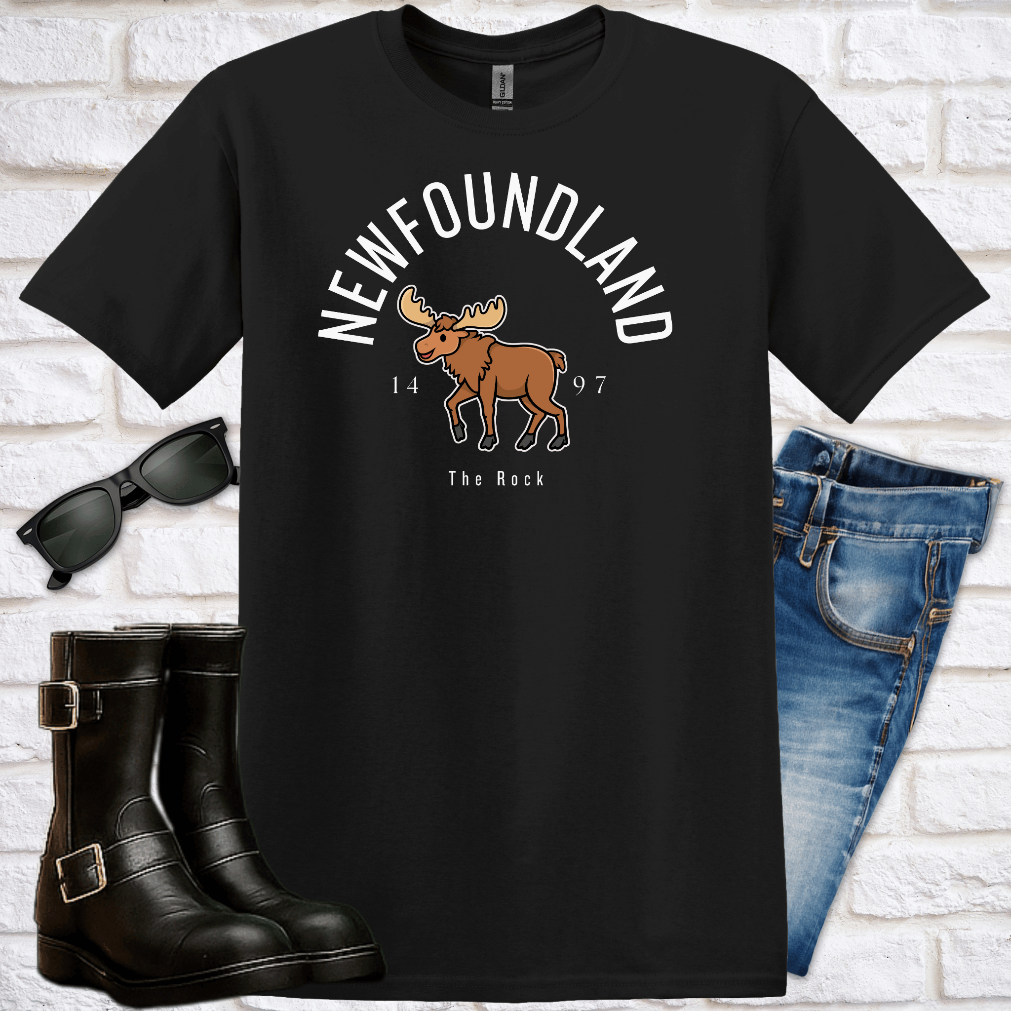 Black t-shirt with moose graphic and text saying Newfoundland, 1497, The Rock, sunglasses, boots, and jeans on a white brick background