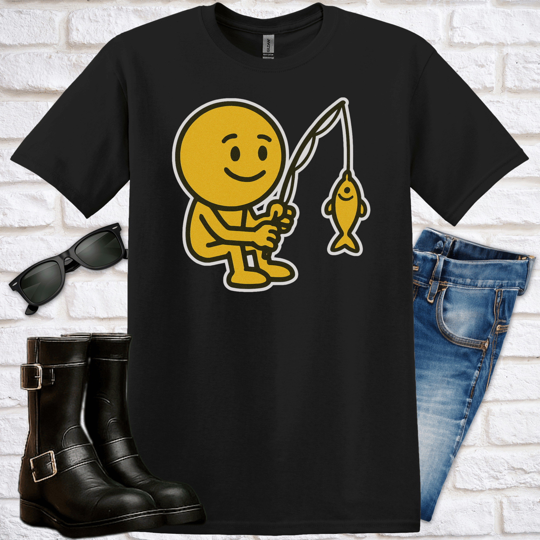 Black t-shirt with yellow graphic of a smiley face emoji fishing, sunglasses, boots, and jeans on a white brick background.