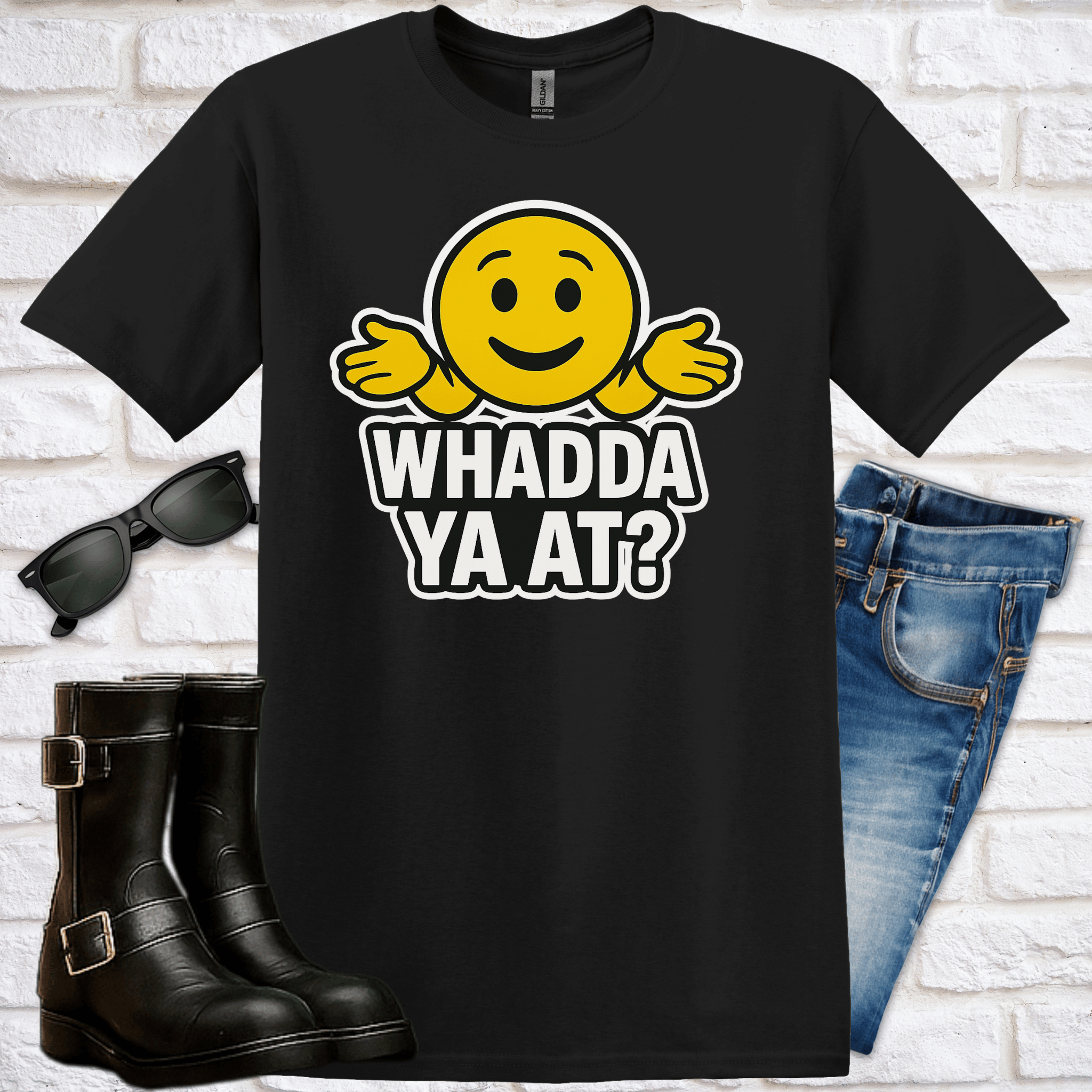 Black t-shirt with yellow emoji face and text saying "Whadda ya at?" on a white brick background with boots, jeans and sunglasses.