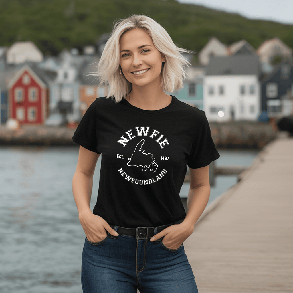 a Woman wearing a black t-shirt with 'NEWFIE' and "Newfoundland" written on it and a map of Newfoundland, standing by a waterfront with houses in the background.