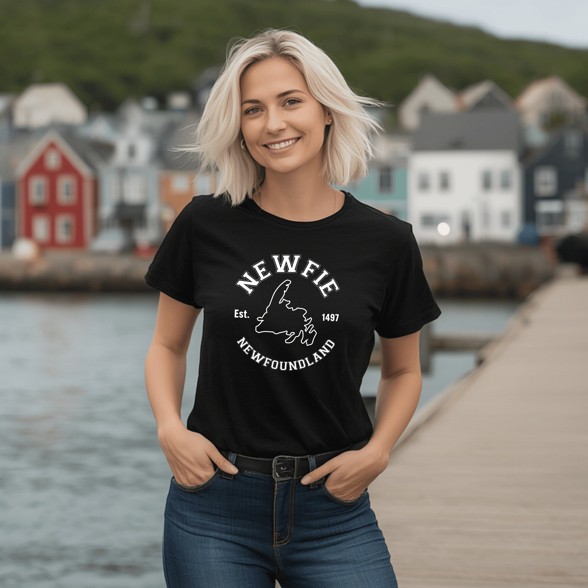 a Woman wearing a black t-shirt with 'NEWFIE' and "Newfoundland" written on it and a map of Newfoundland, standing by a waterfront with houses in the background.