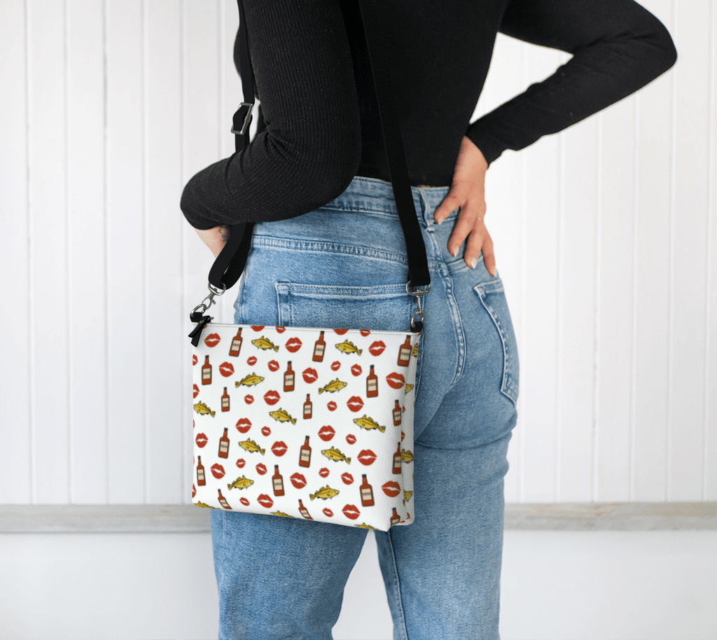 A model wearing a "Screeched In" Crossbody Purse,  featuring a fun pattern of codfish, lipstick kisses, and Screech rum bottles inspired by the iconic island tradition.