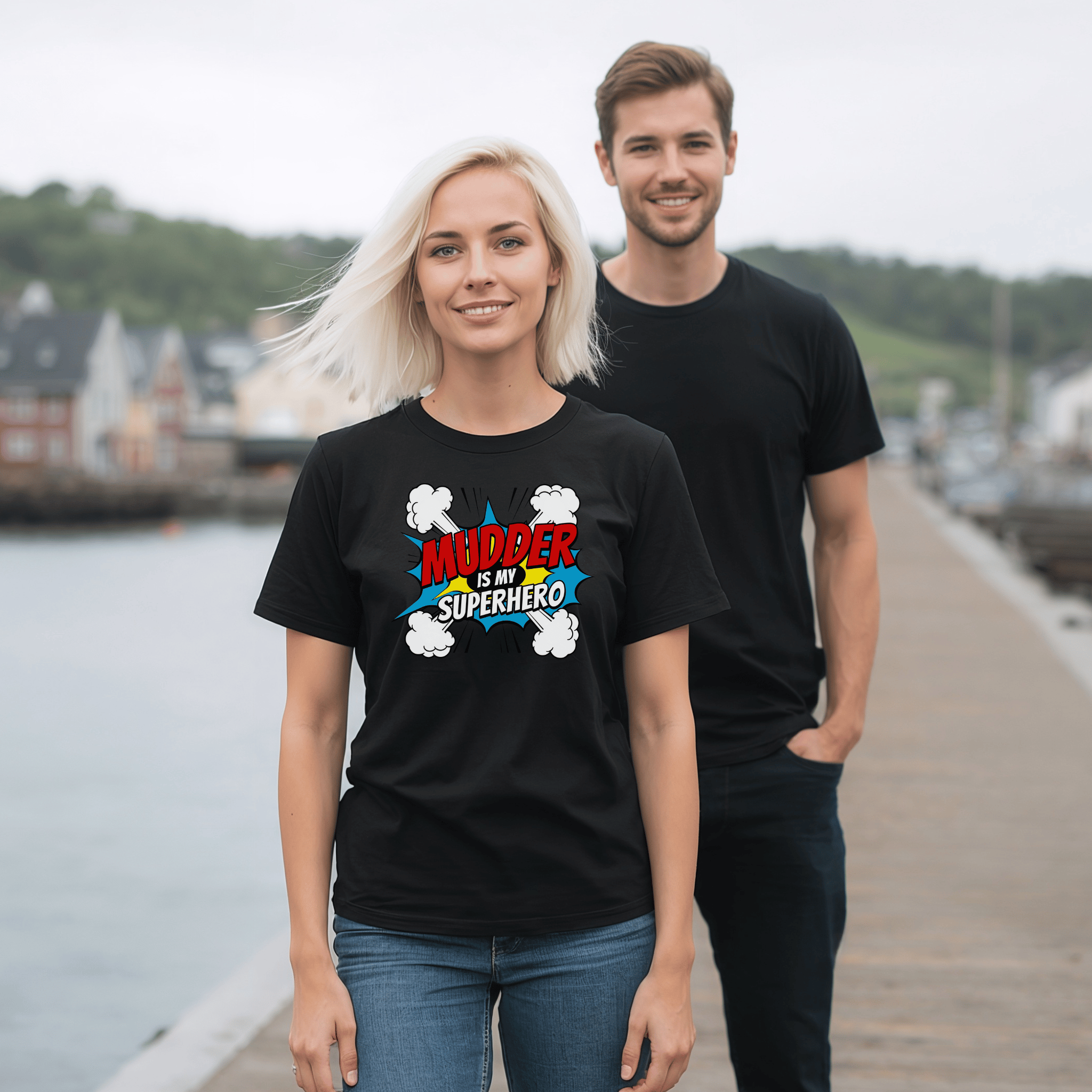 Woman is standing in front of a man, and is wearing a black t-shirts with a "Mudder is my Superhero"  design. They are on a pier. 