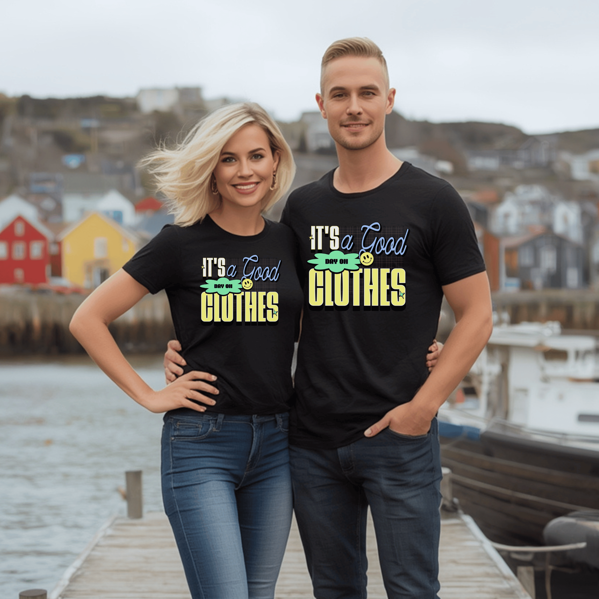 An attractive couple wearing matching black t-shirts with colorful text saying "It's a good day on clothes" with a tiny emoji, standing on a dock with a scenic boat in the background.