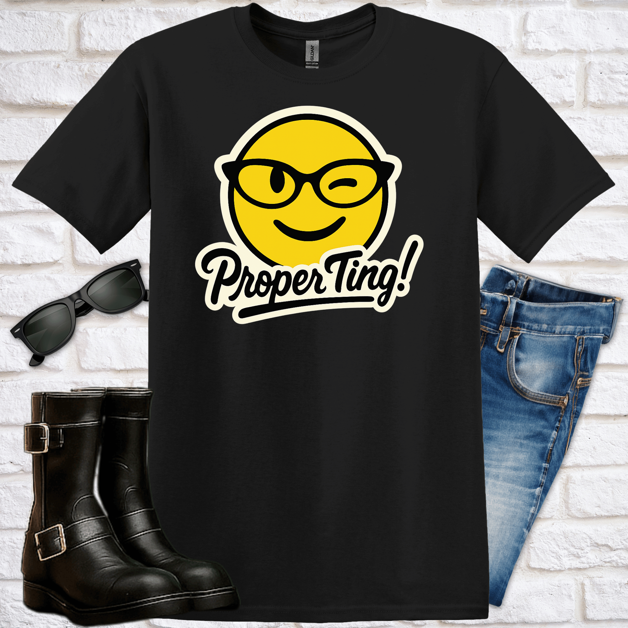 Black t-shirt with yellow smiley face emoji winking from under cat-eyed glasses and 'Proper Ting!' text, paired with sunglasses and jeans on a white brick background.