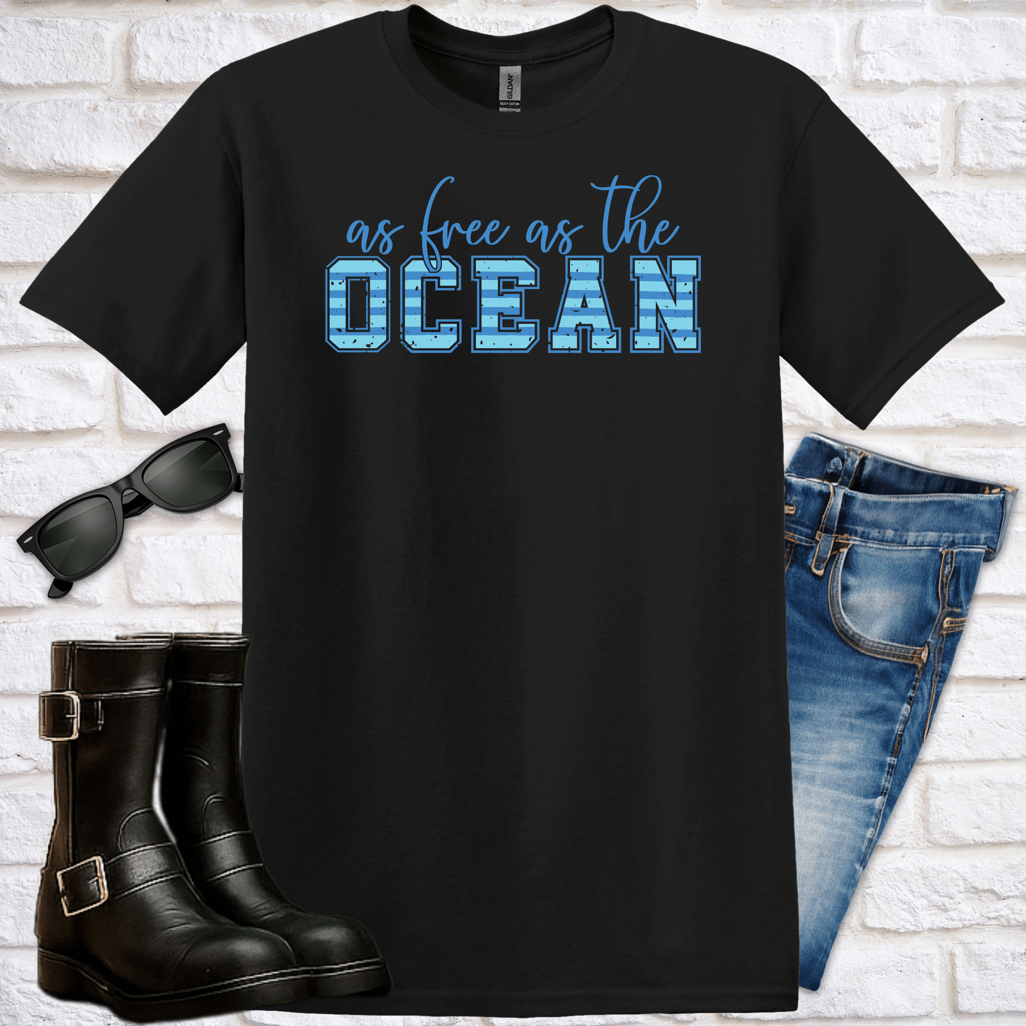Black t-shirt with 'as free as the OCEAN' text, sunglasses, boots, and jeans on a white brick background