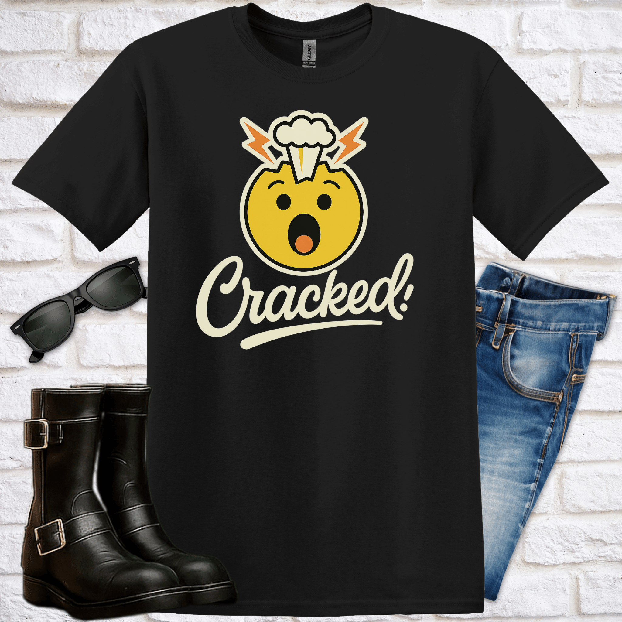 Black t-shirt with a yellow emoji with a funny brain explsion graphic and 'Cracked!' text, sunglasses, jeans  and boots on a white brick background