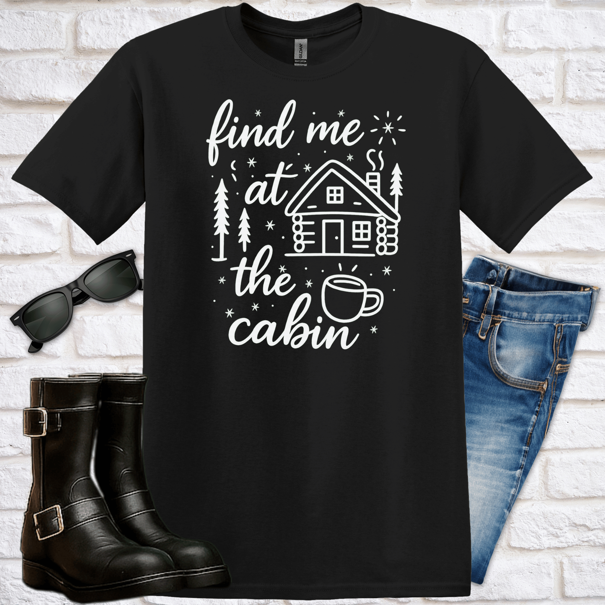 Black t-shirt with 'find me at the cabin' text and a graphic of a cabin and cup of tea or coffee, sunglasses, boots, and jeans on a white brick background