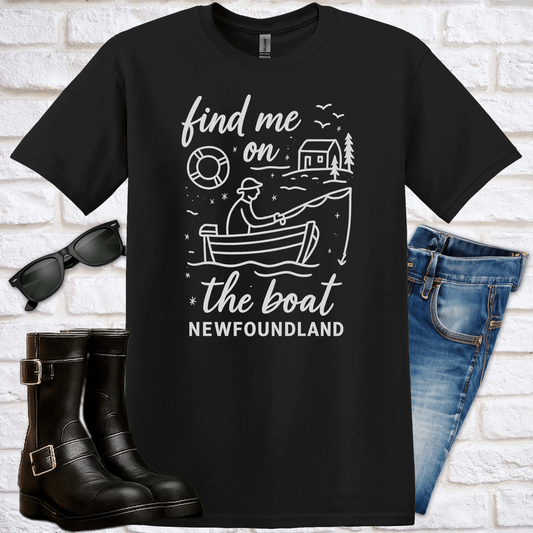 Black t-shirt with a graphic design of a man fishing in a boat and a saltbox house in the background and text saying "find me on the boat, Newfoundland" with sunglasses, boots, and jeans on a white brick background