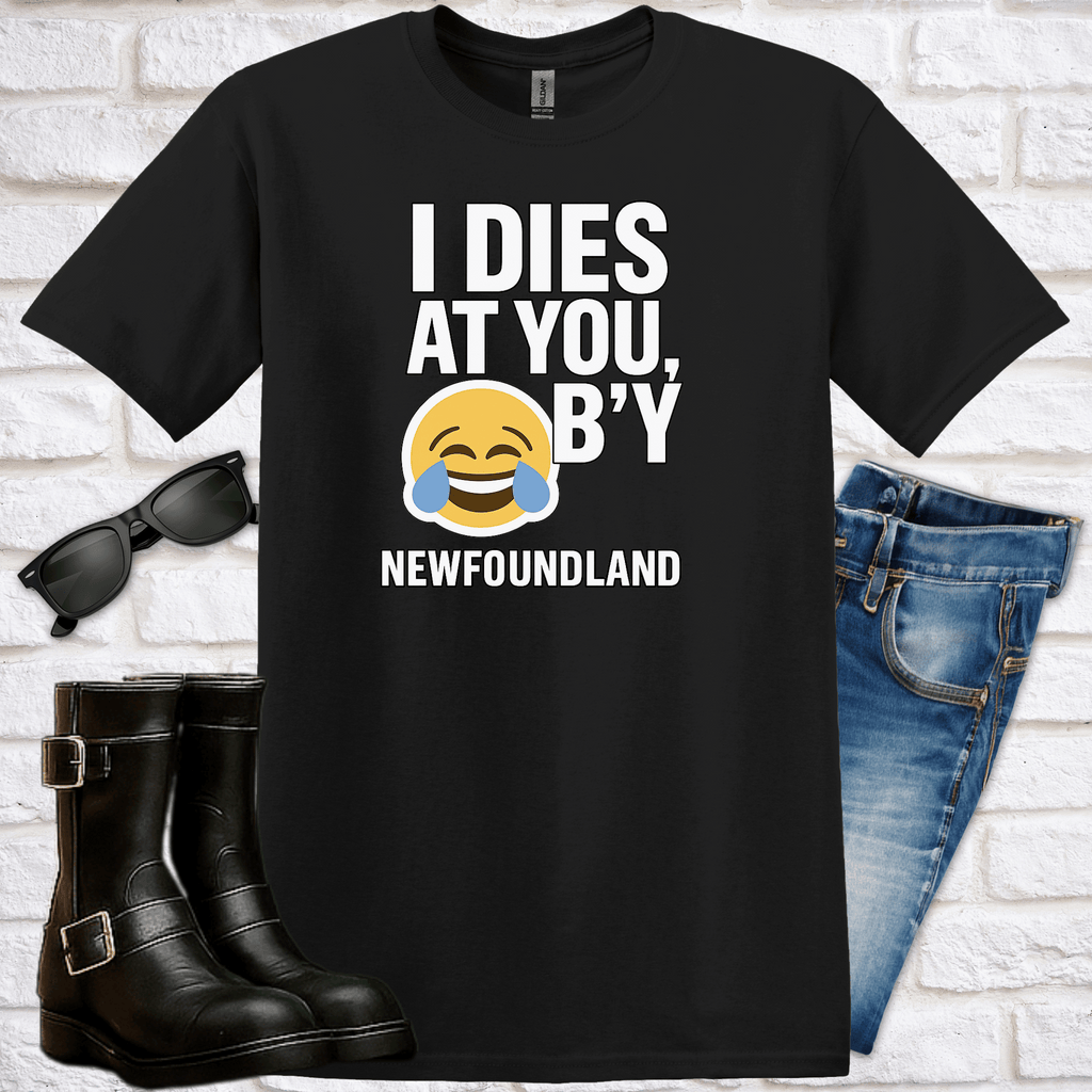 Black t-shirt with humorous text saying "I Dies at You B'y" and an emoji laughing until he is crying, paired with sunglasses, boots, and jeans on a white brick background.