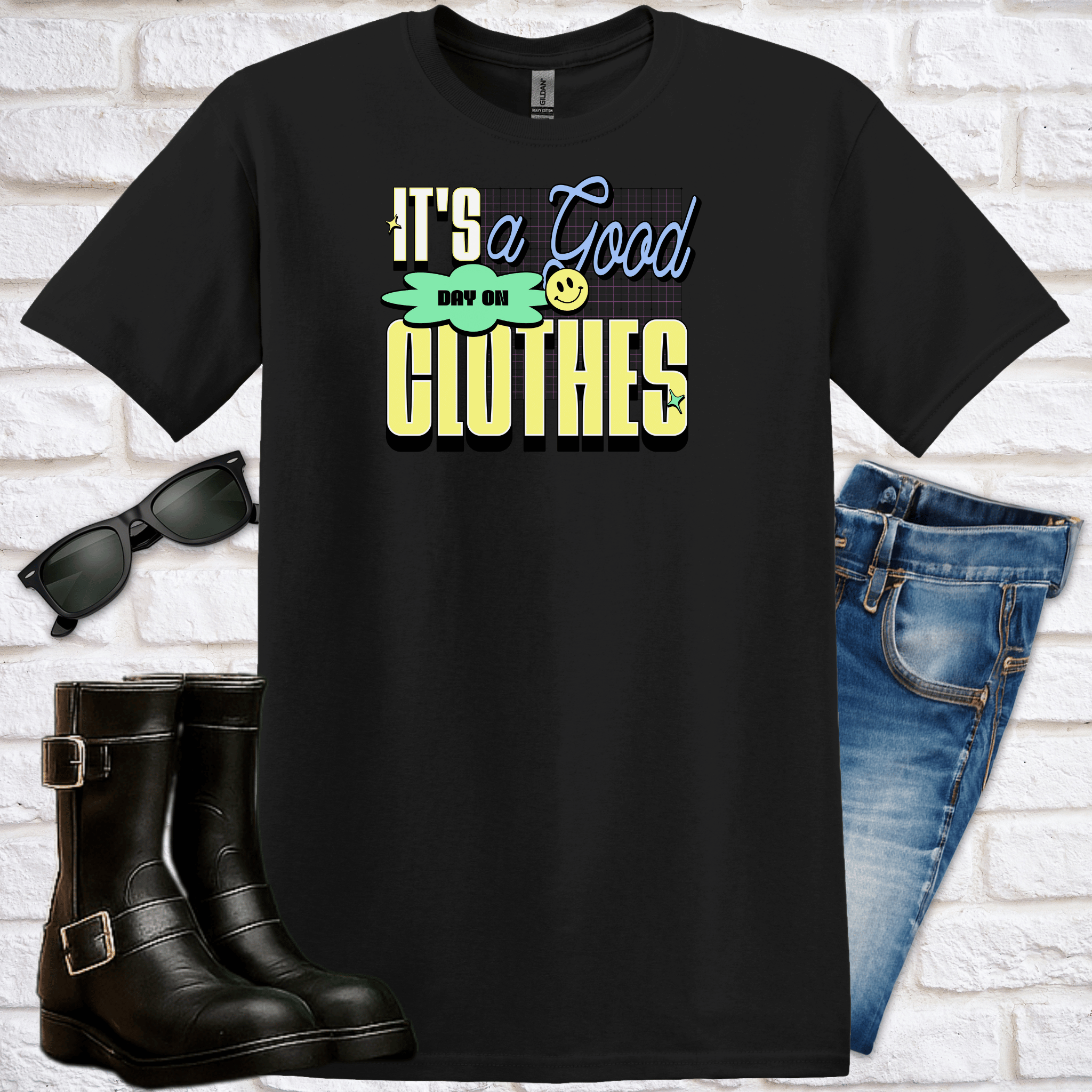 Black t-shirt with colorful text saying "It's a Good Day on Clothes" and a tiny emoji graphic, sunglasses, boots, and jeans on a white brick background