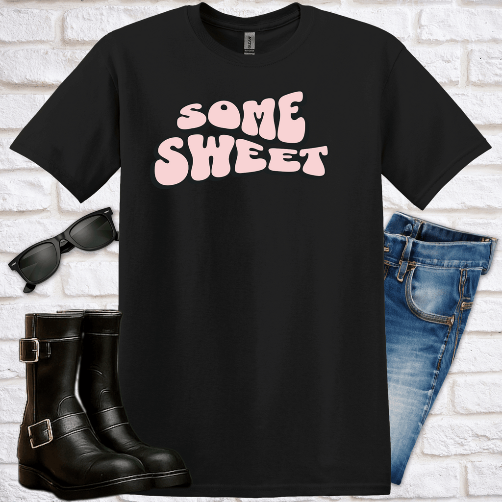 Black t-shirt with 'SOME SWEET' text in a curved, retro style, sunglasses, boots, and jeans on a white brick background
