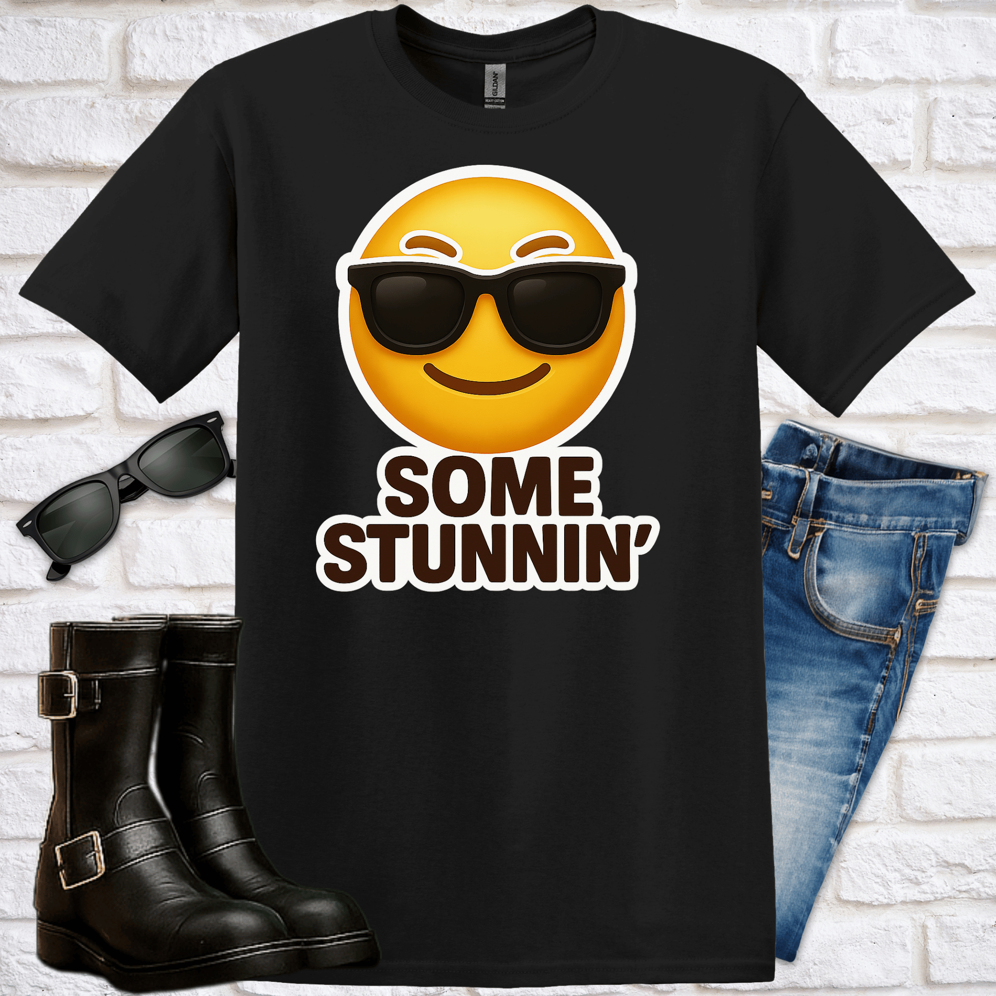 Black t-shirt with emoji and text saying  "Some Stunnin'", sunglasses, boots, and jeans on a white brick background
