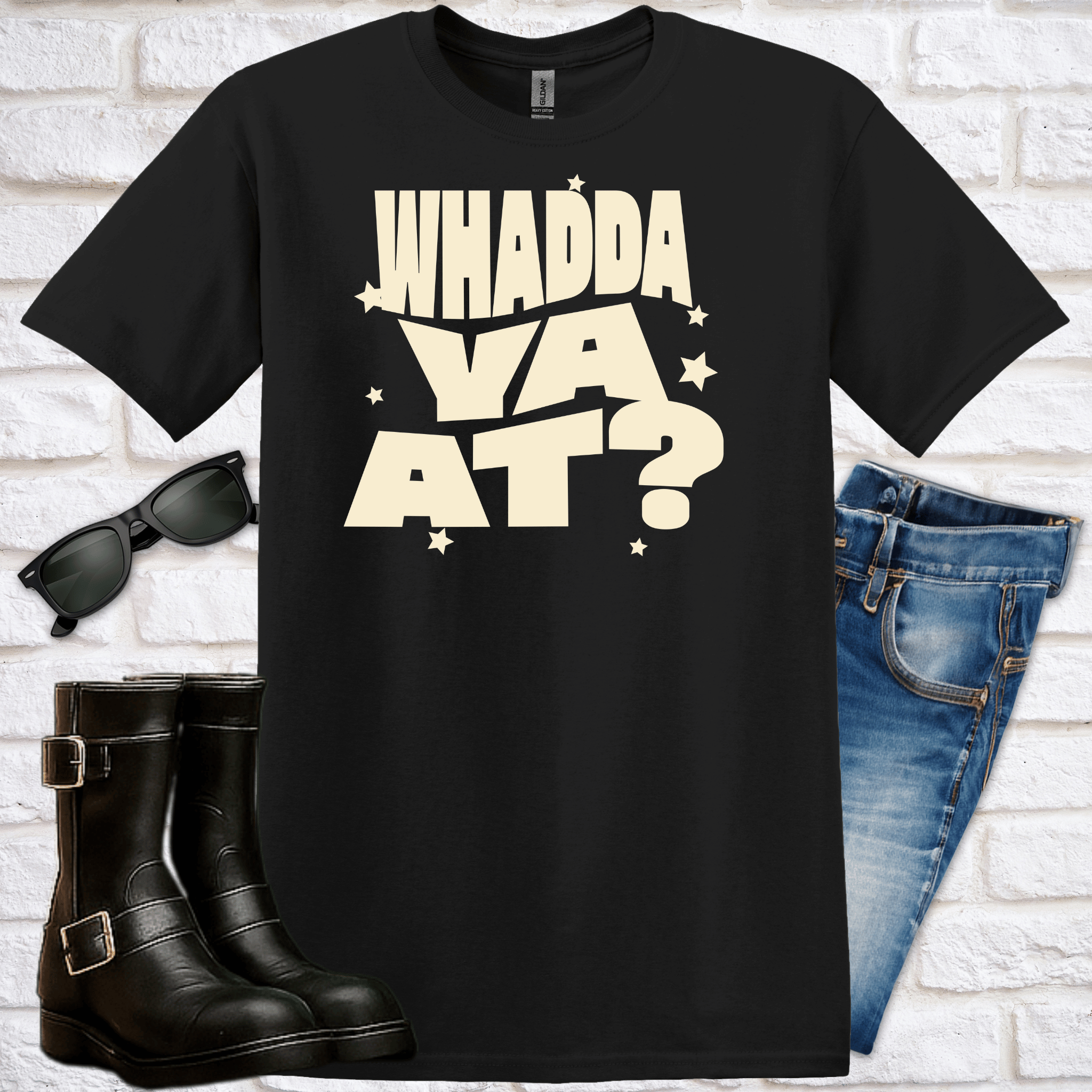 Black t-shirt with cream-colored 'WHADDA VA AT?' text, sunglasses, boots, and jeans on a white brick background