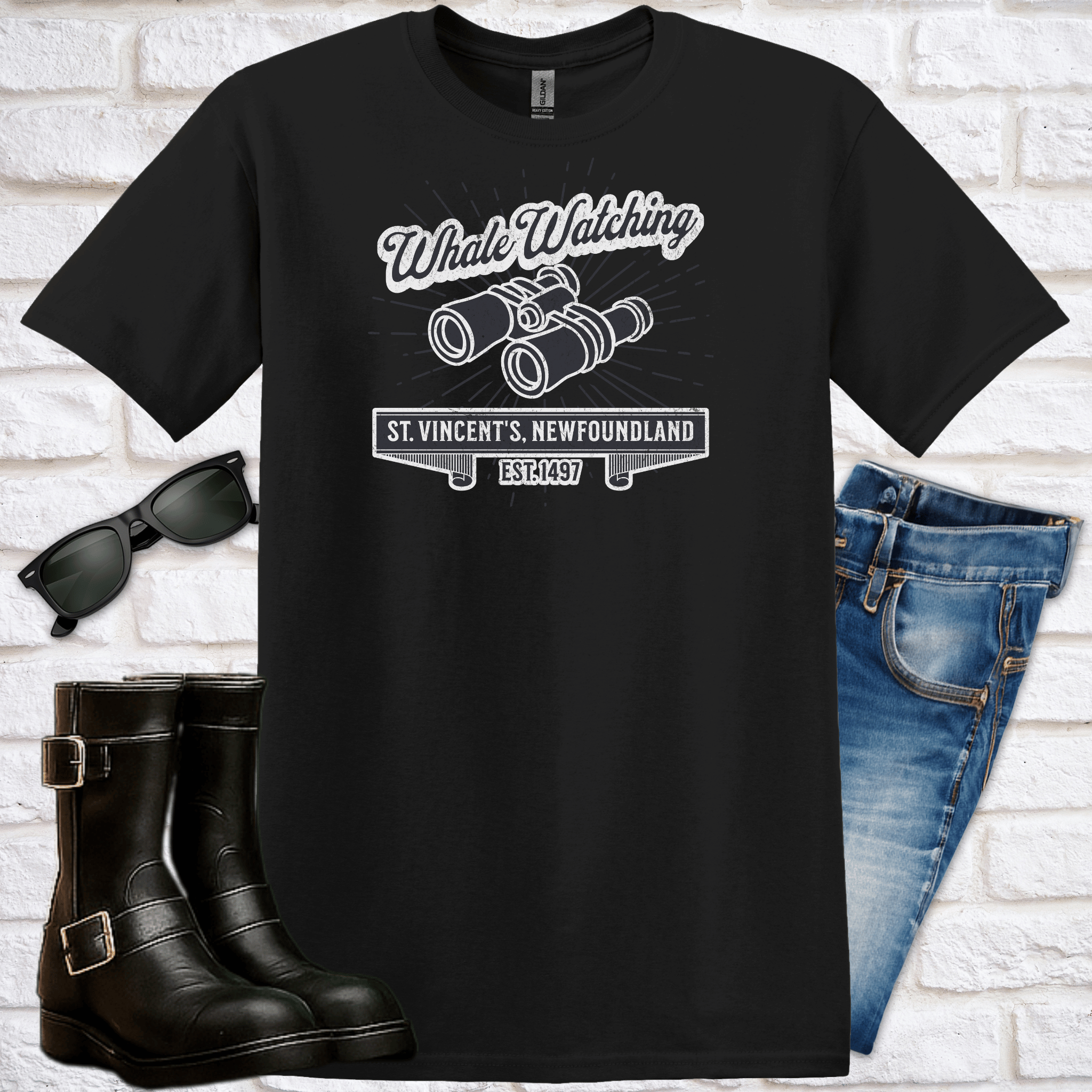 Black t-shirt with 'Whale Watching St. Vincent's Newfoundland' and binoculardesign, sunglasses, boots, and jeans on a white brick background. 