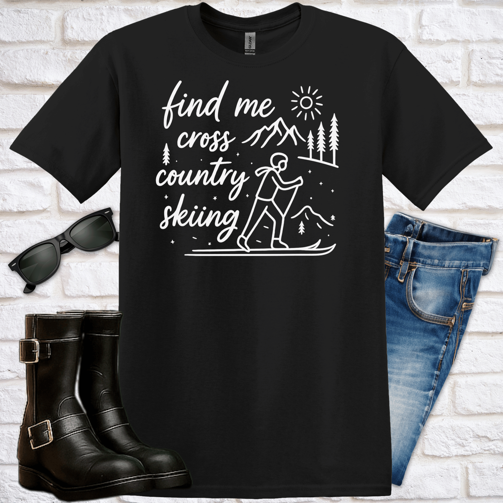 Black t-shirt with cross-country skiing graphic and text saying "find me cross-country skiing", sunglasses, boots, and jeans on a white brick background